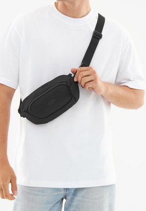Sacoche noire en tissu lisse, présentant une forme incurvée et une poche zippée à l'avant. La sangle réglable permet un port en bandoulière.