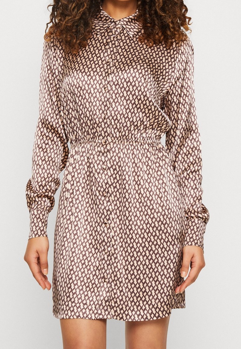 Femme portant une robe mini à manches longues et à boutons, avec un motif géométrique marron et beige, debout devant un fond uni.