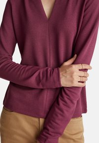 Langärmliges, V-Ausschnitt-Oberteil in Burgunderrot mit strukturierter Strickweise. Der Schnitt ist leger, mit enganliegenden Ärmeln, kombiniert mit beige Hosen.