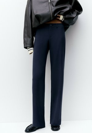 Personne portant un pantalon droit bleu marine taille haute, associé à des mocassins en cuir noirs et une veste courte en cuir noir.