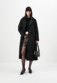 Woman wearing black long quilted coat, black crop top, brown leather skirt, black sheer tights, pointed heeled shoes, and holding black handbag.