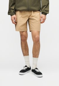 Άνδρας που φοράει μπεζ σορτς, λευκές κάλτσες που φτάνουν μέχρι το μέσο της γάμπας, μαύρα αθλητικά παπούτσια χαμηλού τύπου και πράσινο χακί μπουφάν, στέκεται μπροστά σε ένα απλό φόντο.