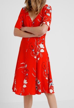 Vrouw draagt een knie-lange rode jurk met witte en zwarte bloemmotieven, korte mouwen en een V-hals, staat met gekruiste armen.