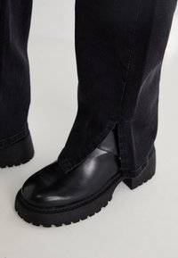 Bota de cuero con suela gruesa negra, llevada con pantalones rectos negros que presentan una pequeña abertura lateral en el dobladillo, sobre un suelo claro.