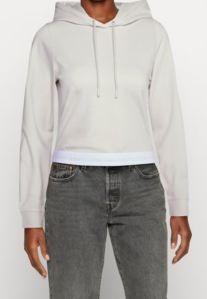 Sweatshirt accorciato grigio chiaro con cappuccio e cordini, caratterizzato da una banda con il logo bianco di Calvin Klein Jeans all'orlo. Indossato con jeans grigio scuro.