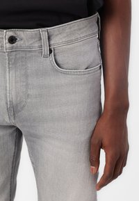 Gray denim jeans with a smooth texture, five-pocket design, black button closure, and contrasting stitching showcased alongside a hand.