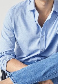 Vaaleansininen nappikauluspaita pehmeästä kankaasta, jossa on klassinen kaulus ja käännetyt hihat, yhdistettynä sinisiin farkkuihin.