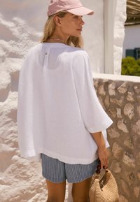 Valge linane särk laiaque varrukatega ja taga nuppe detailiga, koos siniste pinstripe lühikeste pükstega. Hoides põimitud kotti. Pehmed, looduslikud tekstuurid.