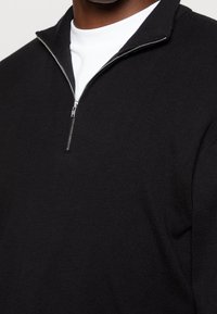 Pull noir à col zippé avec une texture côtelée. Il possède une fermeture éclair argentée avec un col montant, superposé à un t-shirt blanc à col rond.