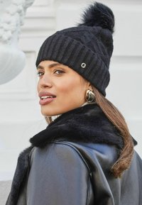Berretto di lana nera a maglia con texture a coste e pom-pom in pelliccia sulla parte superiore, con un piccolo dettaglio del logo. Indossato con una giacca nera con collo in pelliccia sintetica.