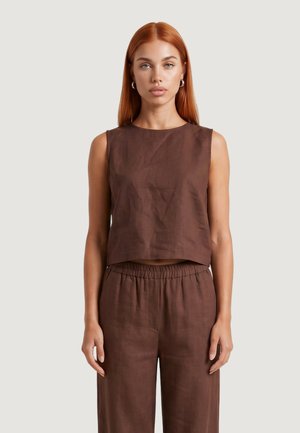 Donna con capelli rossi lisci indossa un top marrone senza maniche e pantaloni larghi abbinati, in piedi davanti a uno sfondo chiaro e semplice.