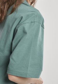 Personne portant un T-shirt vert ample à manches courtes, montrant l'épaule et le bras avec une texture de tissu visible et des détails de couture.
