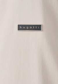 Helles beiges Gewebe mit einer rechteckigen schwarzen Logo-Plakette, auf der das Wort "bugatti" in weißen Buchstaben geprägt ist. Glatte Textur, subtiler Glanz.
