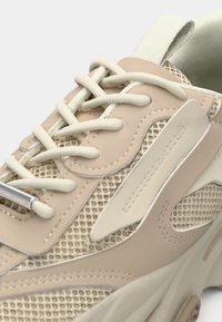 Beige sneaker with mesh panels and smooth accents. Features cream laces and a textured sole. Includes a tab on the heel for easy wear.
