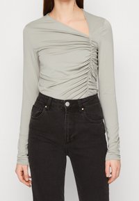 Top a maniche lunghe di colore grigio chiaro con dettagli arricciati sui lati, scollatura a V e tessuto morbido ed elasticizzato abbinato a jeans neri a vita alta.