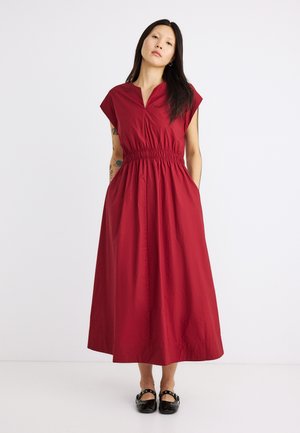 Γυναίκα με μακριά μαύρα μαλλιά, που φοράει κόκκινο midi φόρεμα χωρίς μανίκια με σούρα στη μέση και μαύρα παπούτσια Mary Jane, στέκεται μπροστά σε λευκό φόντο.