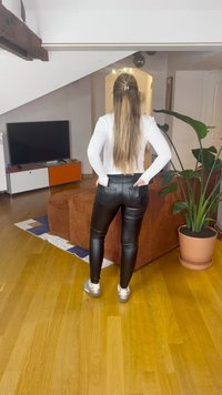 Μαύρα κολάν από δερματίνη με στενή εφαρμογή, δύο πίσω τσέπες, σε συνδυασμό με λευκό μακρυμάνικο τοπ και ασημένια αθλητικά παπούτσια σε σκηνικό σαλονιού.