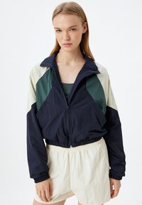 Vetoketjullinen takki, jossa on tummansinisen, vihreän ja kermaisen värin blokkeja; elastiset hihansuut ja helma; kevyt kangas; yhdistetty kermaisiin shortseihin.