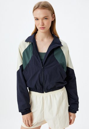Zip-up jas met marineblauwe, groene en crèmekleurige kleurblokken; elastische manchetten en zoom; lichtgewicht stof; gecombineerd met crèmekleurige shorts.
