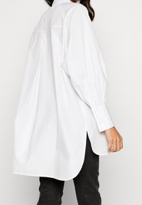 Camicia bianca a maniche lunghe con colletto a punta, dettagli delle maniche arricciate e spacchi laterali per facilitare i movimenti. Tessuto di cotone morbido.