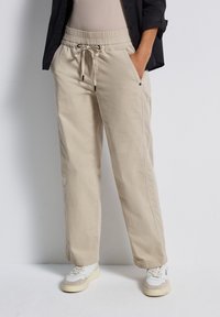 Pantalon léger extensible beige avec une taille élastique et un cordon de serrage. Il possède des poches latérales et une coupe décontractée, associé à des baskets blanches.