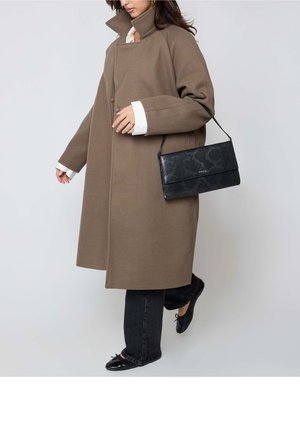 Vrouw met een oversized bruine jas, zwarte jeans, zwarte ballerina's, zichtbare witte manchetknopen, en een zwarte schoudertas met textuur.