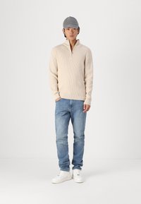 Beżowy sweter z dzianiny na zamek, niebieskie dżinsy, białe trampki i szara czapka, stojący na tle białego, gładkiego tła.