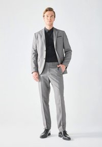 Costum gri din țesătură netedă, cu sacou cu un singur rând de nasturi și pantaloni cu față plată, asortat cu o cămașă neagră cu nasturi și pantofi negri.