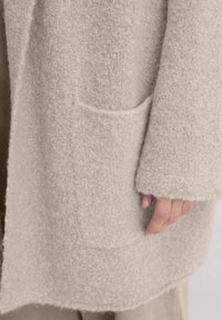 Beige, fuzzy gebreid vest met een patchzak, verlaagde schouders en brede mouwen. De textuur is zacht, met een licht onregelmatig oppervlak.