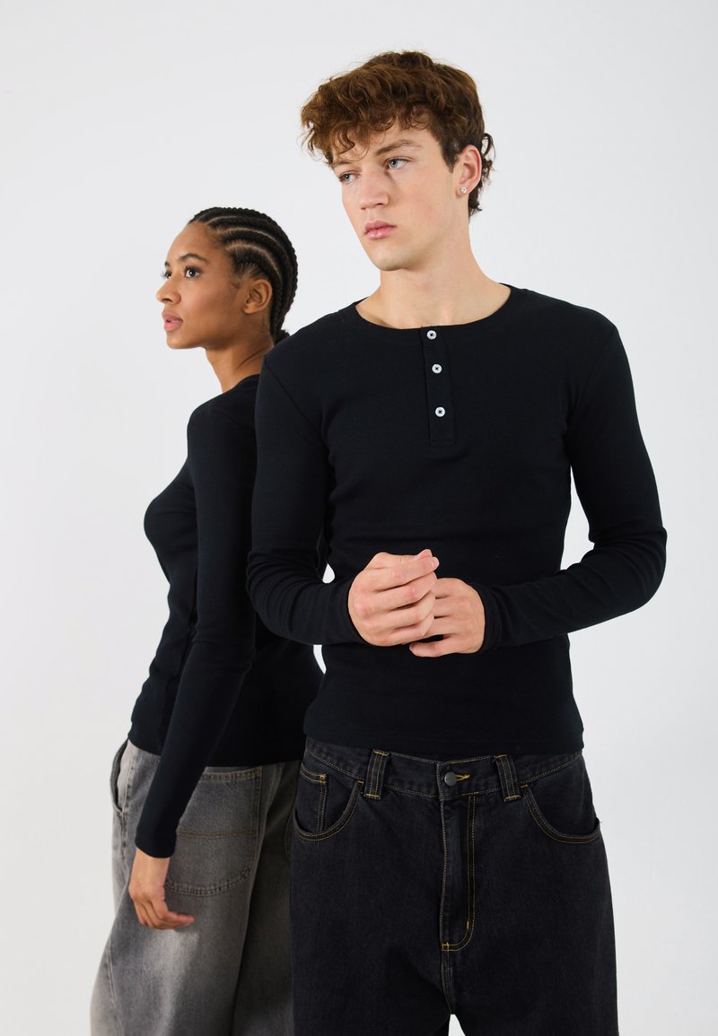 Črne dolge majice henley imajo gumbno zapiranje spredaj in so v kombinaciji z ohlapnimi črnimi in sivimi kavbojkami. Gladek material, prilegajoča se zasnova.