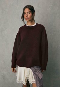 Burgundy oversized striktrøje med rund halsudskæring, der har en blød tekstur. Lagd med en hvid kjole med blonder og stribet stof.