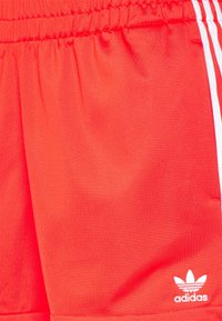 Červené sportovní šortky s texturovaným povrchem, vybavené elastickým pasem a bílými bočními pruhy. Obsahují malé bílé logo.