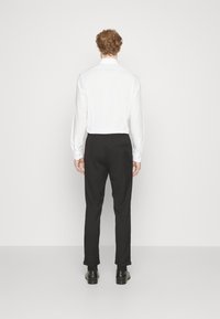 Chemise blanche à manches longues, pantalon noir et chaussures noires, vue de dos. La chemise a un col standard ; le pantalon possède une poche arrière.