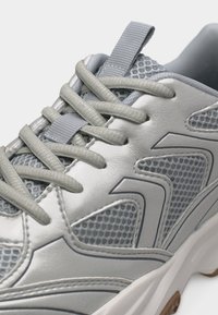 Γκρι αθλητικό παπούτσι με πάνελ από δίχτυ, χοντρές κορδόνια δεμένα σε κανονικό σταυρωτό σχέδιο και χοντρή λευκή σόλα με καφέ λεπτομέρειες.