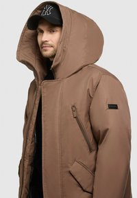 Braune gepolsterte Jacke mit großer Kapuze, seitlichem Reißverschlussfach und einem Logo-Patch am Ärmel. Glatte Textur, minimalistisches Design.