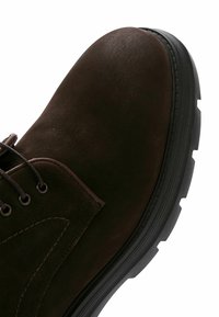 Chaussure en cuir marron avec une tige texturée, bout rond et semelle en caoutchouc noir présentant des motifs de relief profonds. L'œillet pour les lacets est visible.