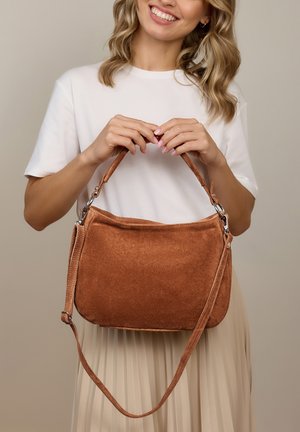 Mujer sonriente con camisa blanca sostiene un bolso de ante marrón con asa superior y una correa larga sobre un fondo liso.