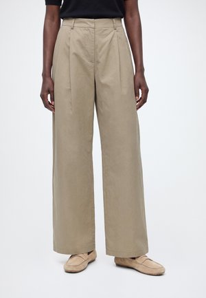 Personne portant un pantalon beige taille haute à jambes larges et des mocassins beige clair, debout sur un fond blanc uni.