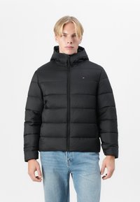 Jachetă puffer neagră cu glugă, având secțiuni orizontale cusute, fermoar frontal și un mic logo pe pieptul stâng.