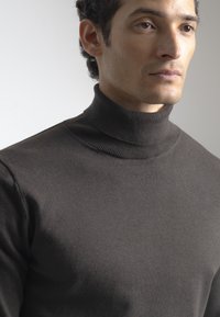 Hombre vistiendo un suéter de cuello alto de canalé marrón oscuro, mirando hacia la derecha con una expresión neutral sobre un fondo gris claro.