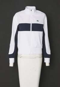 Chaqueta blanca con cremallera y raya horizontal azul marino. Presenta un cuello alto, puños elásticos y el logo de Lacoste en el pecho. Textura de tela suave.