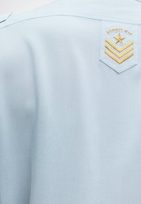 Ανοιχτό μπλε ύφασμα με ραμμένο έμβλημα ασπίδας που απεικονίζει ένα χρυσό αστέρα, τρεις σήματα και το κείμενο "SCHOTT NYC" στο πάνω μέρος της πλάτης.