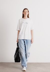 Witte katoenen t-shirt met "Calvin Klein Since 1968" in het grijs gedrukt, gecombineerd met lichtblauwe denim jeans en een zwarte tote bag.