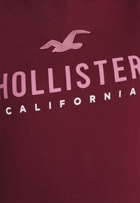 Camiseta de algodón burdeos con un logo de gaviota bordado en rosa y texto en blanco "HOLLISTER CALIFORNIA" en una fuente sans-serif en negrita.