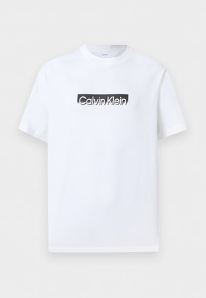 Valge lühikeste varrukatega ümarakaelusega T-särk, millel on must ristkülikukujuline logomärk ja esiküljel tsentreeritud "Calvin Klein" tekst.