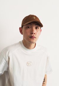 Brown cap with a curved brim, featuring a small heart emblem. Paired with a white sporty t-shirt displaying a beige Nike logo.