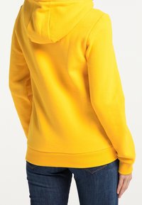 Sweat-shirt jaune à capuche, tissu texturé, poche kangourou, poignets et ourlet côtelés. Porté avec un jean bleu.