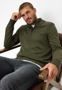 Armeegrün Pullover mit hohem Kragen und Viertelreißverschluss. Rippbündchen und Saum, über einem weißen Hemd getragen. Das Modell sitzt auf einem Stuhl.