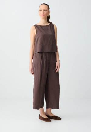 Kobieta stojąca w brązowej, bezrękawnikowej bluzce w prążki i szerokich spodniach, ubrana w pasujące brązowe baleriny, na jednolitym jasnym tle.