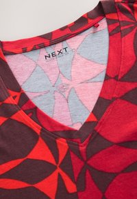 Red and dark brown slouchy V-neck tee featuring a geometric floral pattern, made from soft fabric with a smooth texture. Brand label visible.